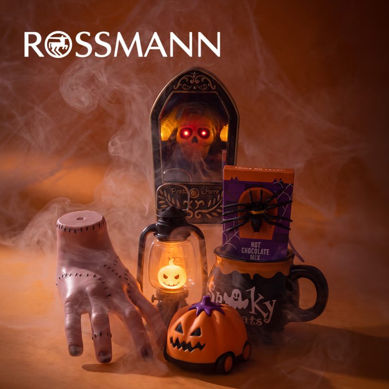 Dekoracje na Halloween na pomarańczowym tle z dymem: sztuczna ręka ze szwami, dzwonek z czaszką o świecących czerwonych oczach, latarnia z podświetlaną dynią, pojazd w kształcie dyni oraz kubek z opakowaniem czekolady do picia ozdobionym pająkiem. U góry białe logo Rossmann.